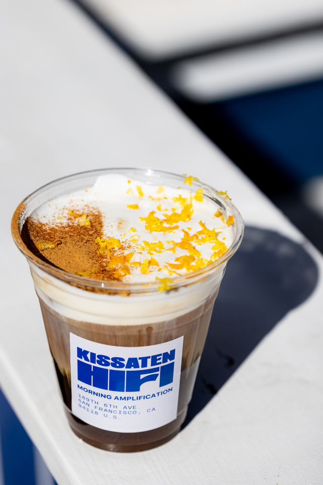A clear plastic cup filled with layered iced coffee topped with foam, sprinkled cinnamon, and orange zest, labeled “Kissaten Hi-Fi Morning Amplification.”